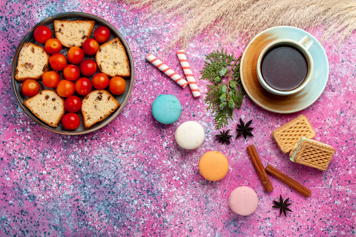 Top view delicious sliced cake with sour fresh plums french macarons and tea on the pink desk
