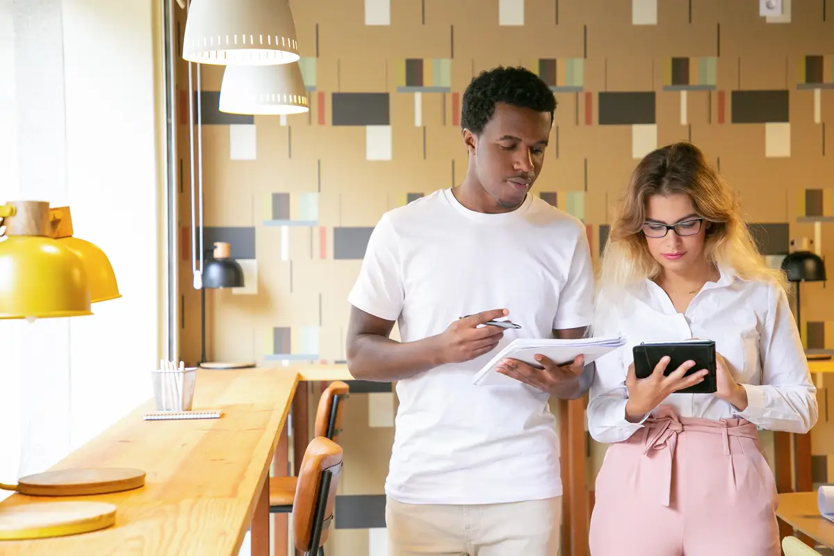 Diverse colleagues discussing project in coworking office, using tablet together, writing notes in notebook while standing at workplaces and talking