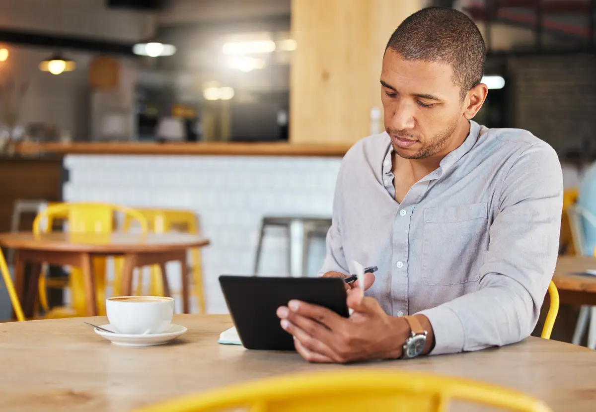 Serious freelance business man working in a coffee shop doing remote work for his startup Young male entrepreneur using a tablet planning growth strategy in a cafe and writing notes