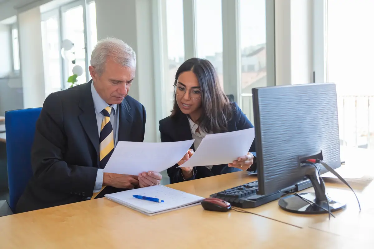 LLC nos EUA: Como Abrir, Vantagens e Diferenças da Corporation 2 Female professional explaining document details to client at workplace. Serious business leader consulting financial or legal expert. Teamwork or cooperation concept