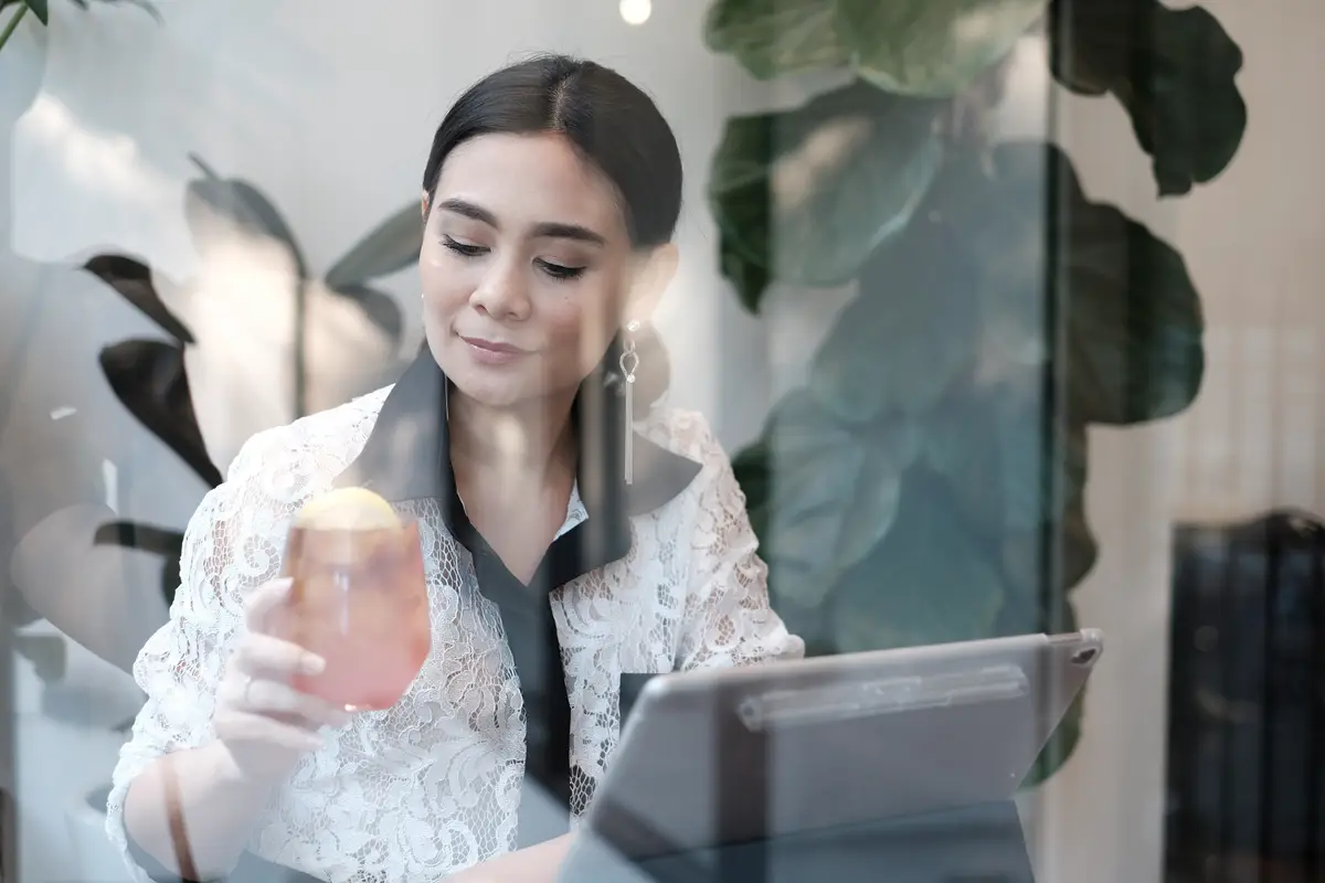 Woman holding drink while using digital tablet seen through glass