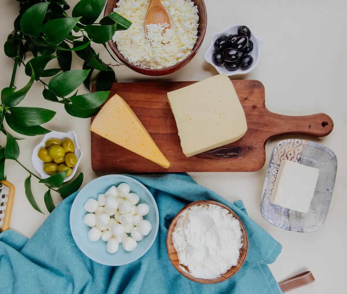 Top view of different kind of cheese on wooden cutting board and cottage cheese in a wooden bowl with pickled olives on rustic table