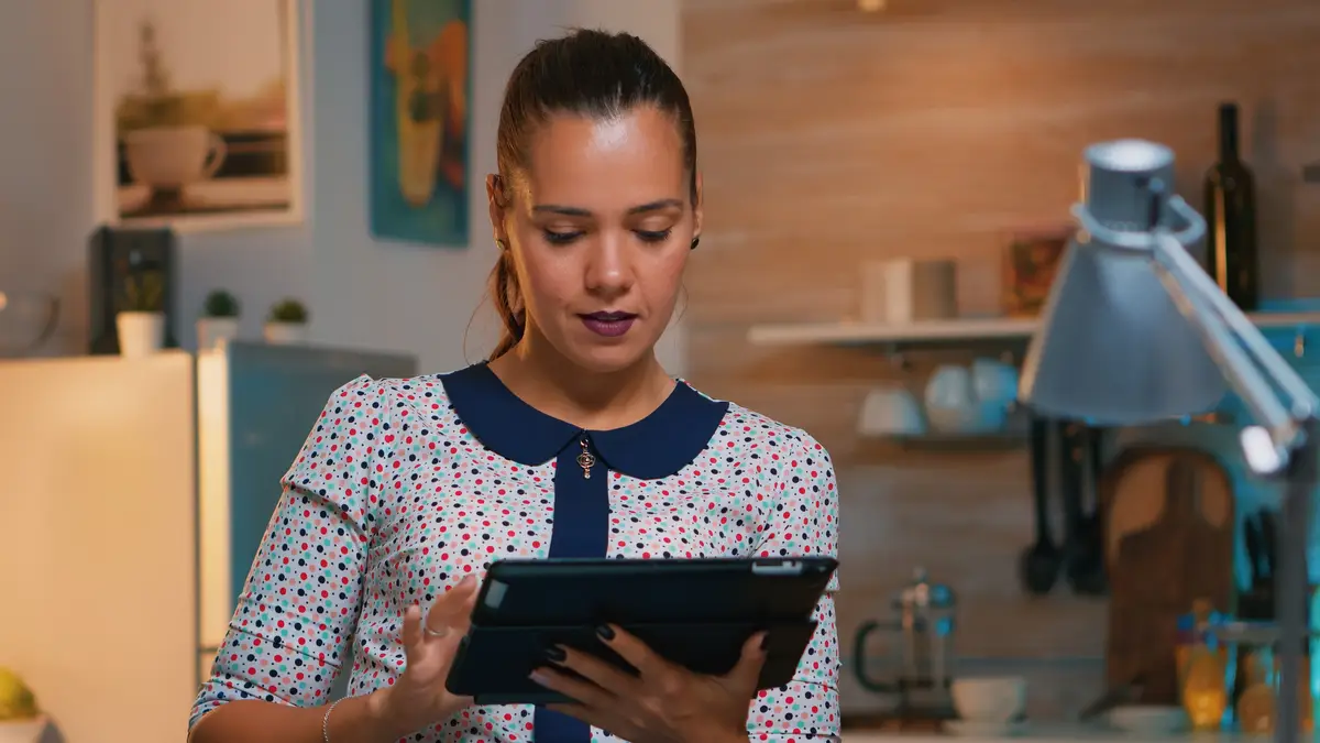 Businesswoman taking break using tablet late at night sitting in modern kitchen. Busy focused employee using modern technology network wireless doing overtime writing, searching.