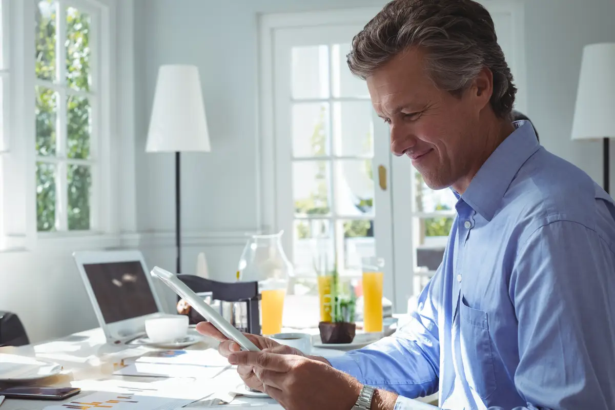 Man in his 40s reviewing tablet at modern home workspace with laptop papers coffee copy space