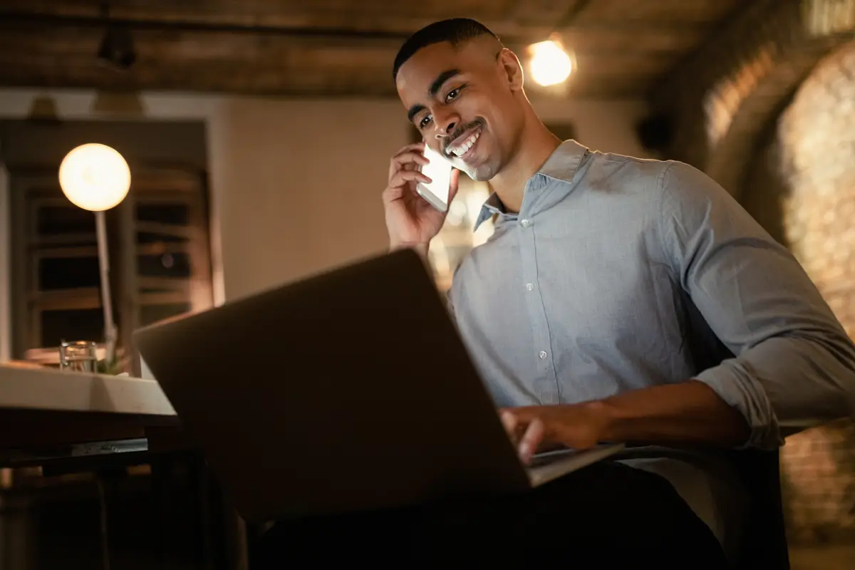 Low angle view of happy African American entrepreneur talking on the phone while working late on laptop in the office