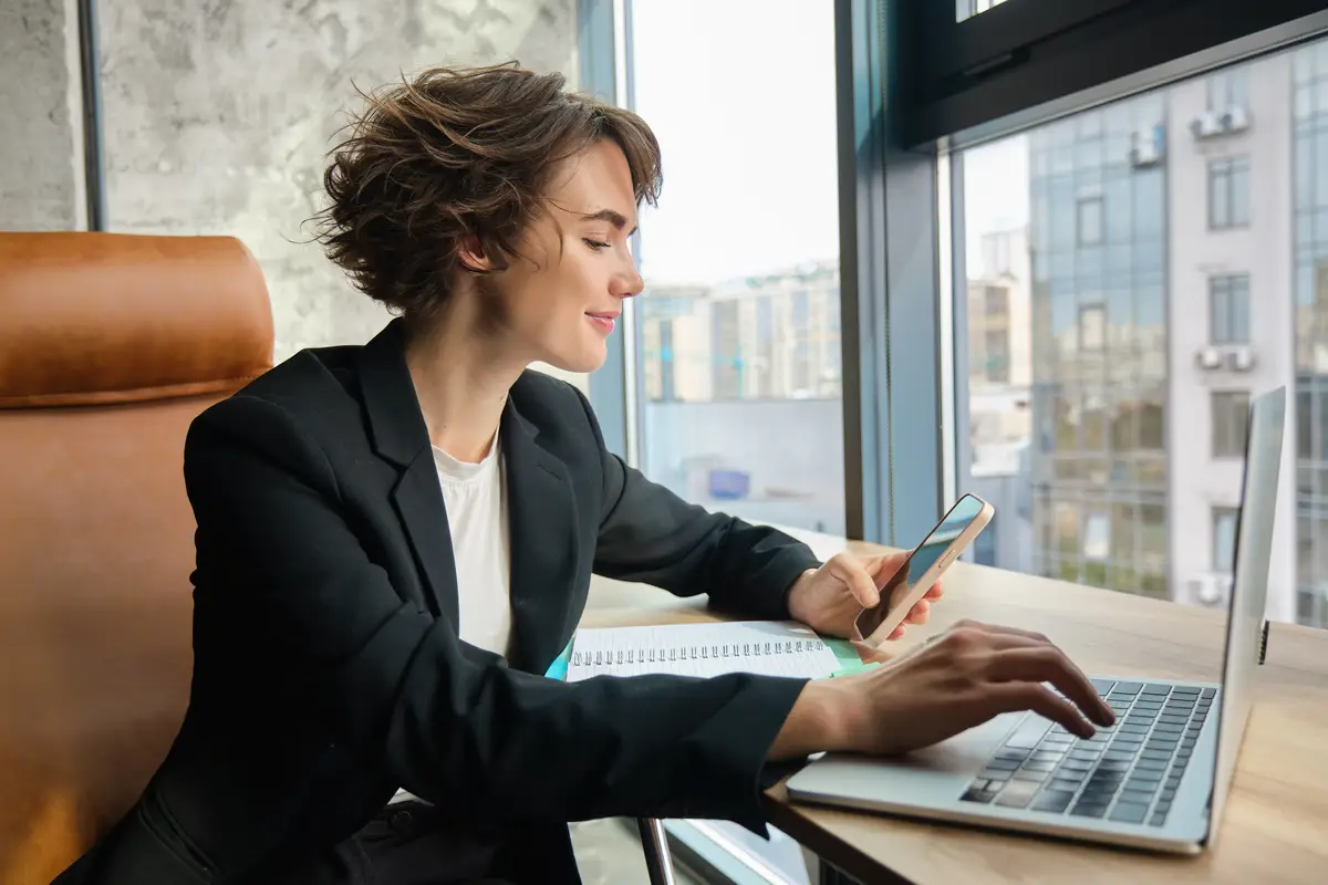 Young working woman in office sitting in front of computer and using smartphone wearing suit