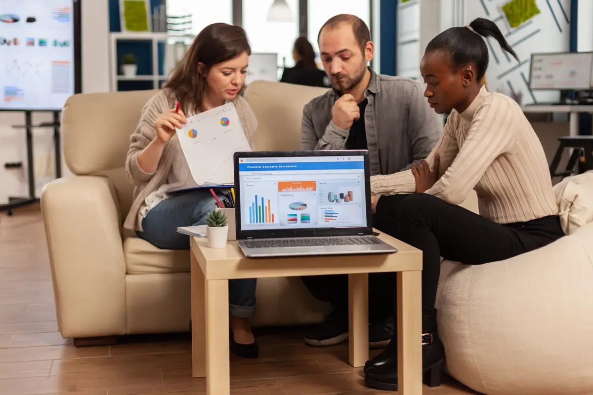 Diverse colleagues employees cooperating in office discussing at workplace, analysing graphs sitting on couch