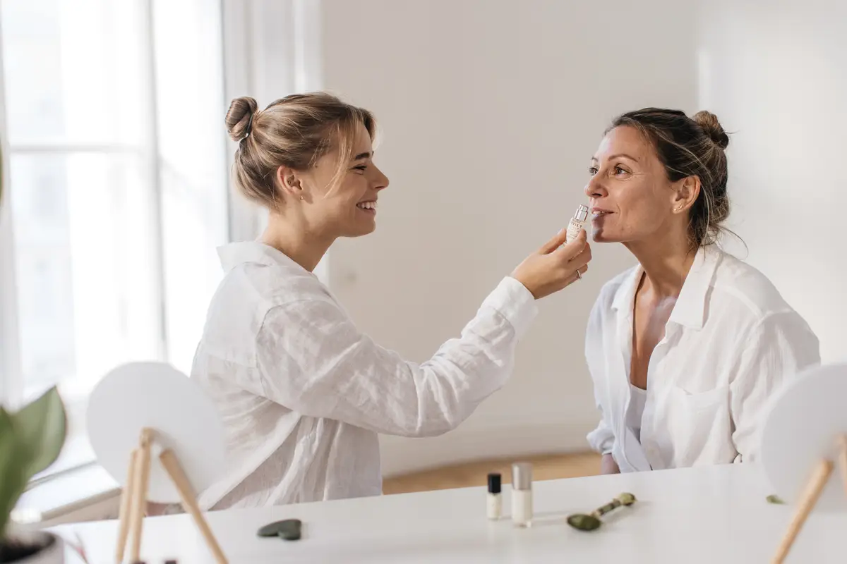 Young caucasian girl gives sniff of facial serum to adult woman sitting in front of her at table indoors. Home spa facial concept