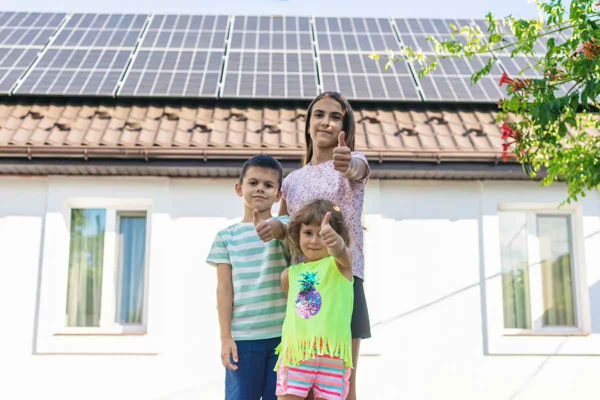 Three smiling children showing thumbs up gesturemeaning classin front of a house with solar panels