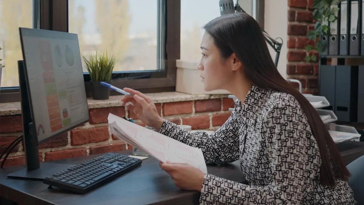 Asian woman comparing charts on papers and computer, working to plan business project in startup office. Company employee analyzing documents to design financial data and strategy.