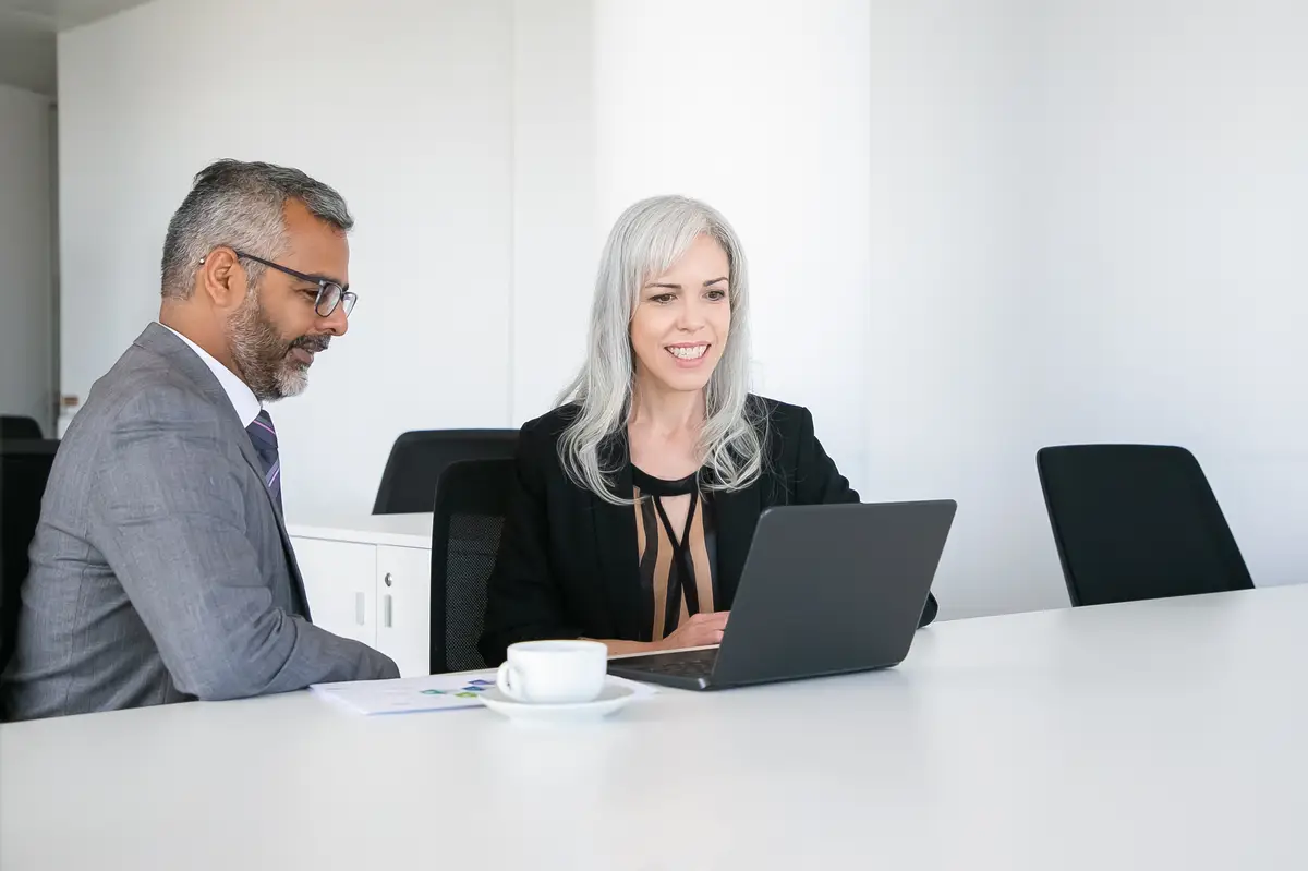 Two happy colleagues using laptop for video call, sitting at table with cup of coffee, looking at display and talking. Online communication concept