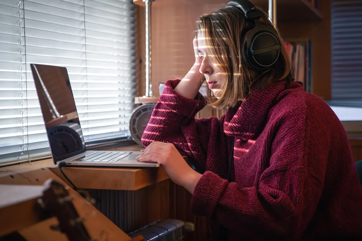 A girl with headphones behind a laptop in the sunlight through the blinds