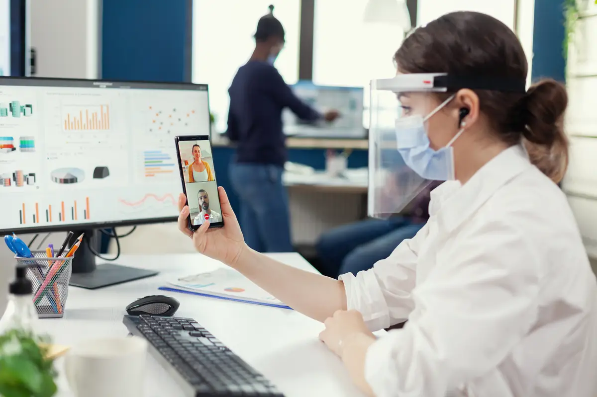 Businesswoman using wireless headphones during online conference on smartphone wearing face mask in workplace. Multiethnic coworkers working respecting social distance in business during global pandem