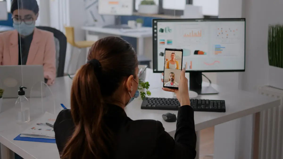 Executive business woman wearing protective face mask using phone for online videocall conference with remotely team. Coworkers working in background respecting social distance