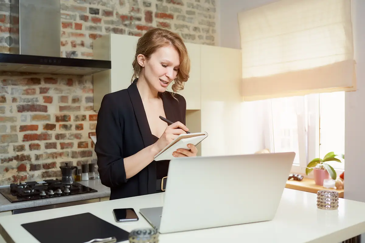 A lady works remotely on a laptop in a kitchen. A girl with a smile doing notes into the notebook during the report of a colleague at a video conference at home.