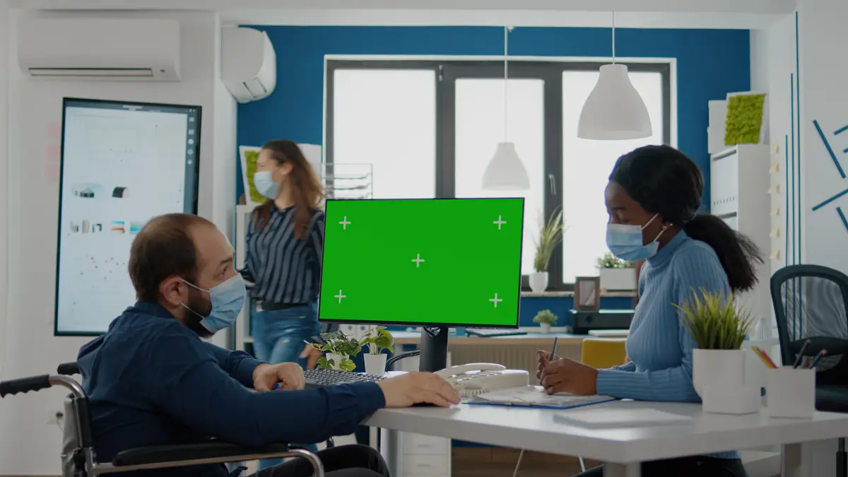 Handicapped paralysed invalid entretreneur with face mask explaning to black employee financial evolution of company pointing at green screen, mock up, chroma key izolated desktop in new normal office