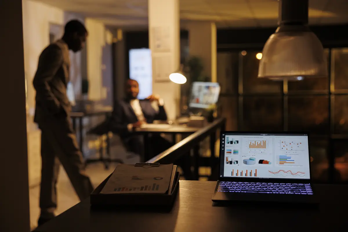 Laptop computer with company statistics standing on desk table in startup office, business team working overhours at marketing strategy in background. Coworkers discussing financial growth report