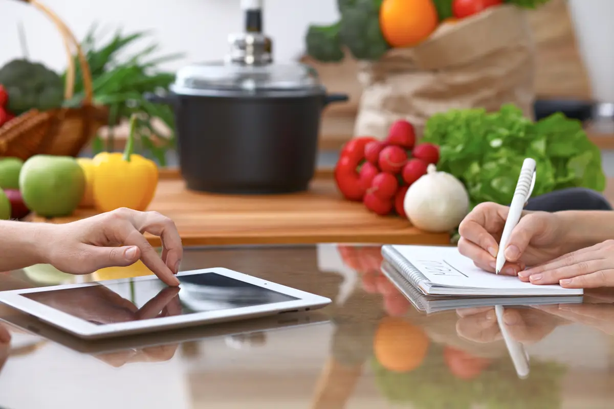 Human hands of two female persons using touchpad for making menu in the kitchen. Closeup of two women are making online shopping by tablet computer and credit card. Cooking and shopping concept.