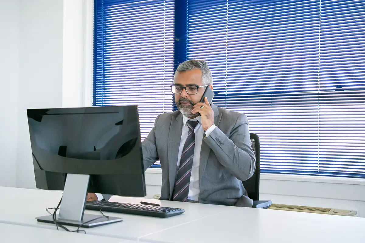 Serious mature business leader speaking on cell phone while using computer at workplace in office. Medium shot. Digital communication and multitasking concept