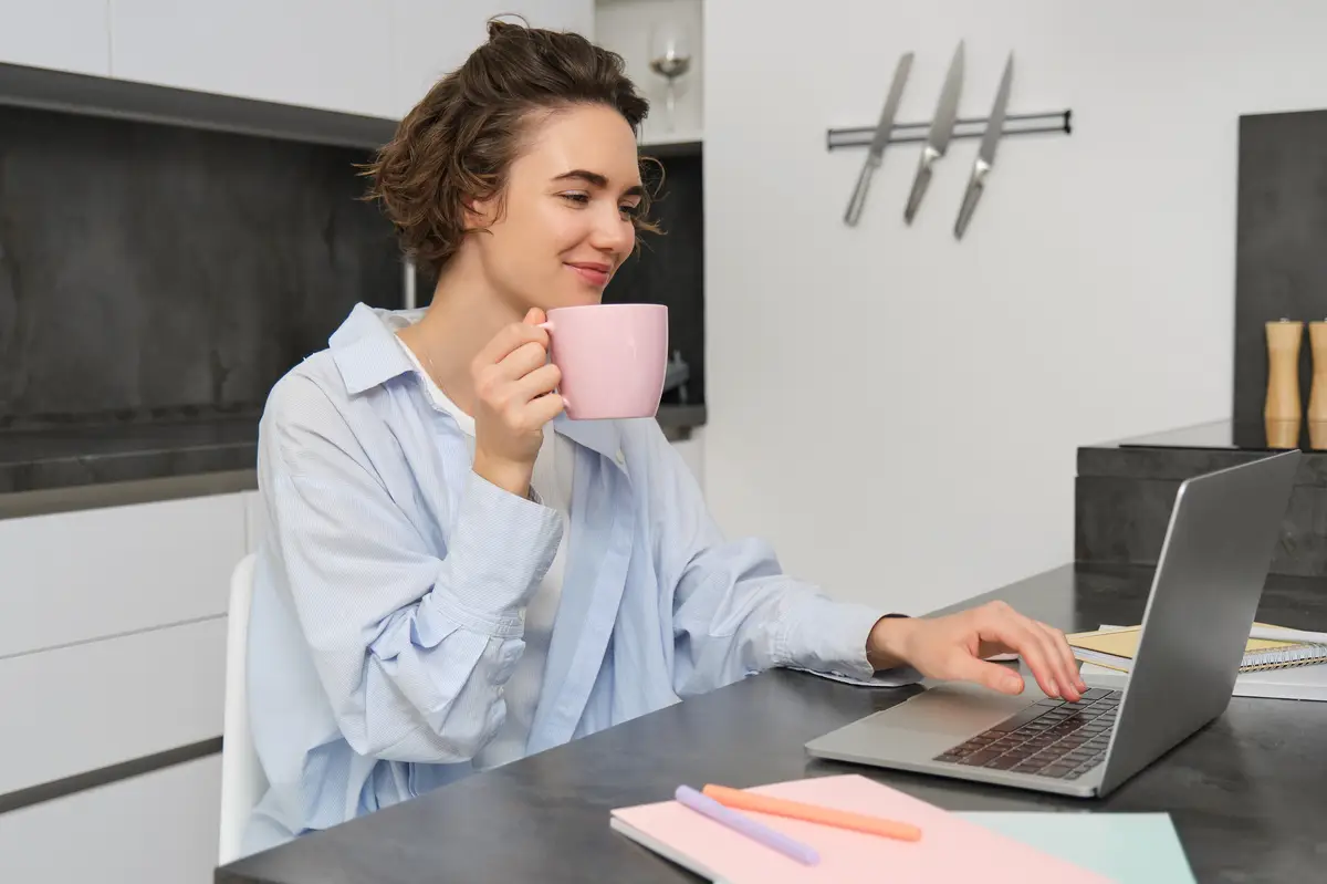 Beautiful girl working from home drinking coffee and looking at laptop Young businesswoman sits in kitchen and manages her business via computer