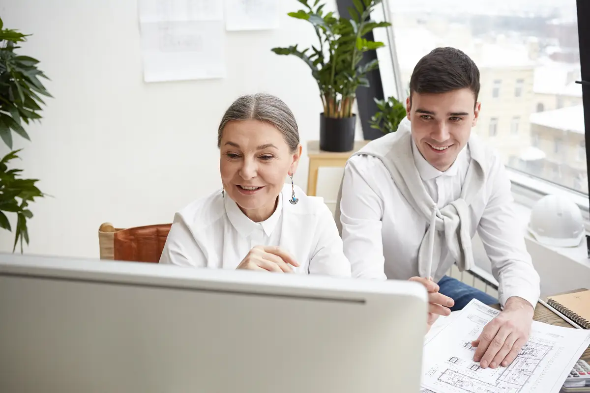 Skilled smiling mature female chief engineer using CAD program on computer while her cheerful young male assistant studying blueprint on desk in front of him. Success, teamwork and collaboration