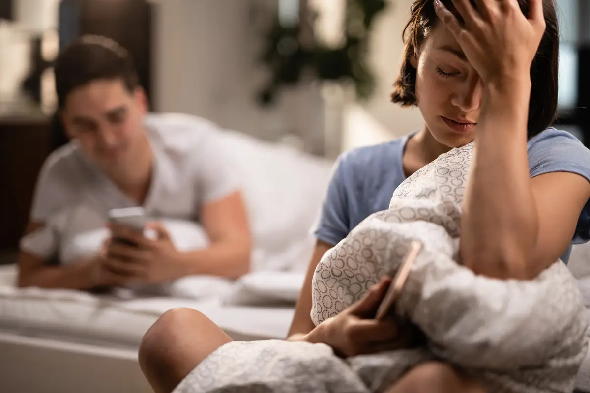 Young sad woman with headache sitting in the bedroom while her boyfriend is text messaging on mobile phone in the background