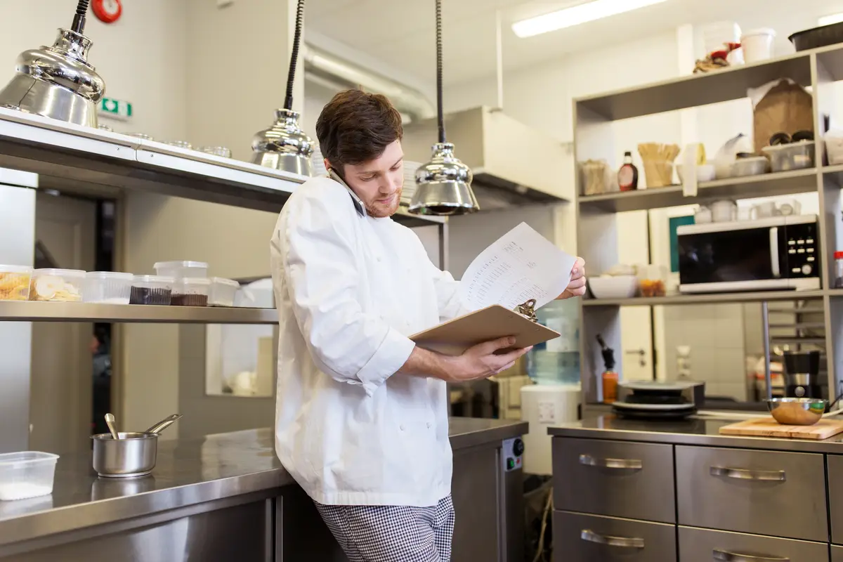 cooking, profession and people concept - male chef cook with clipboard calling on smartphone at restaurant kitchen