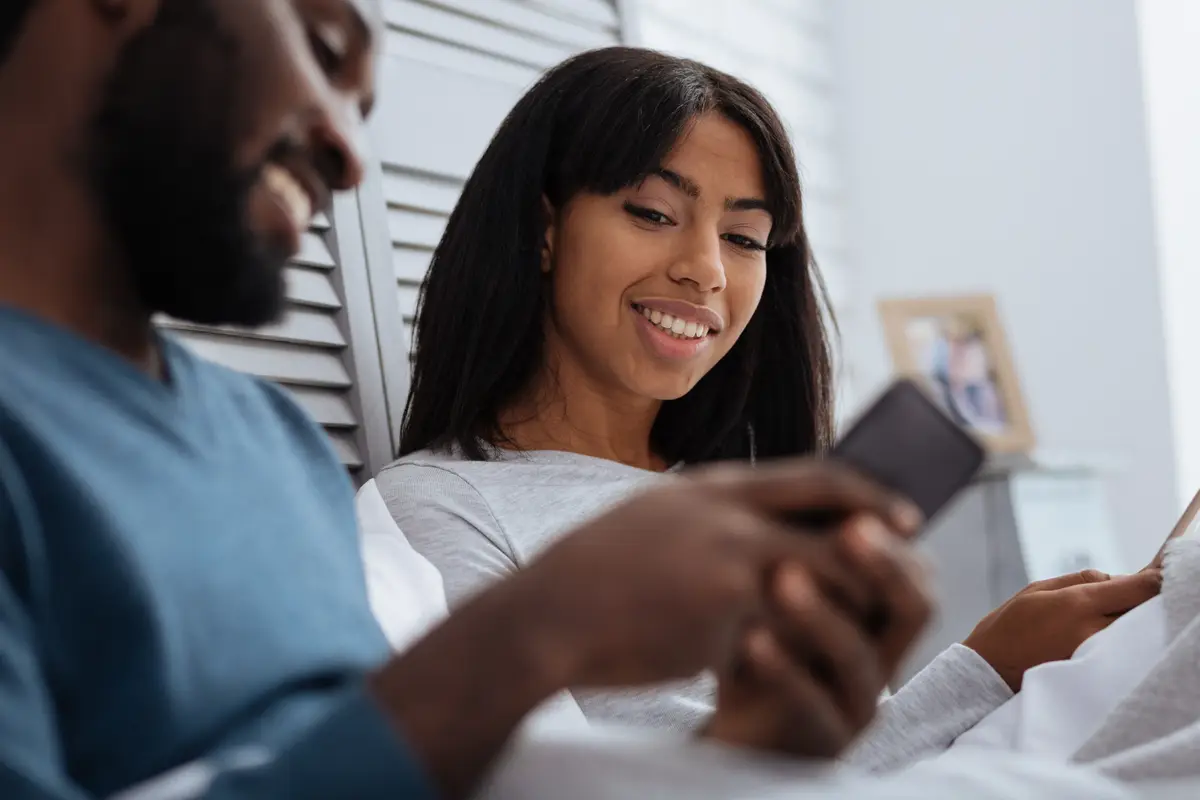 Smiling couple. Happy young smiling people looking at the screen of a smart phone while sitting in their comfortable bedroom