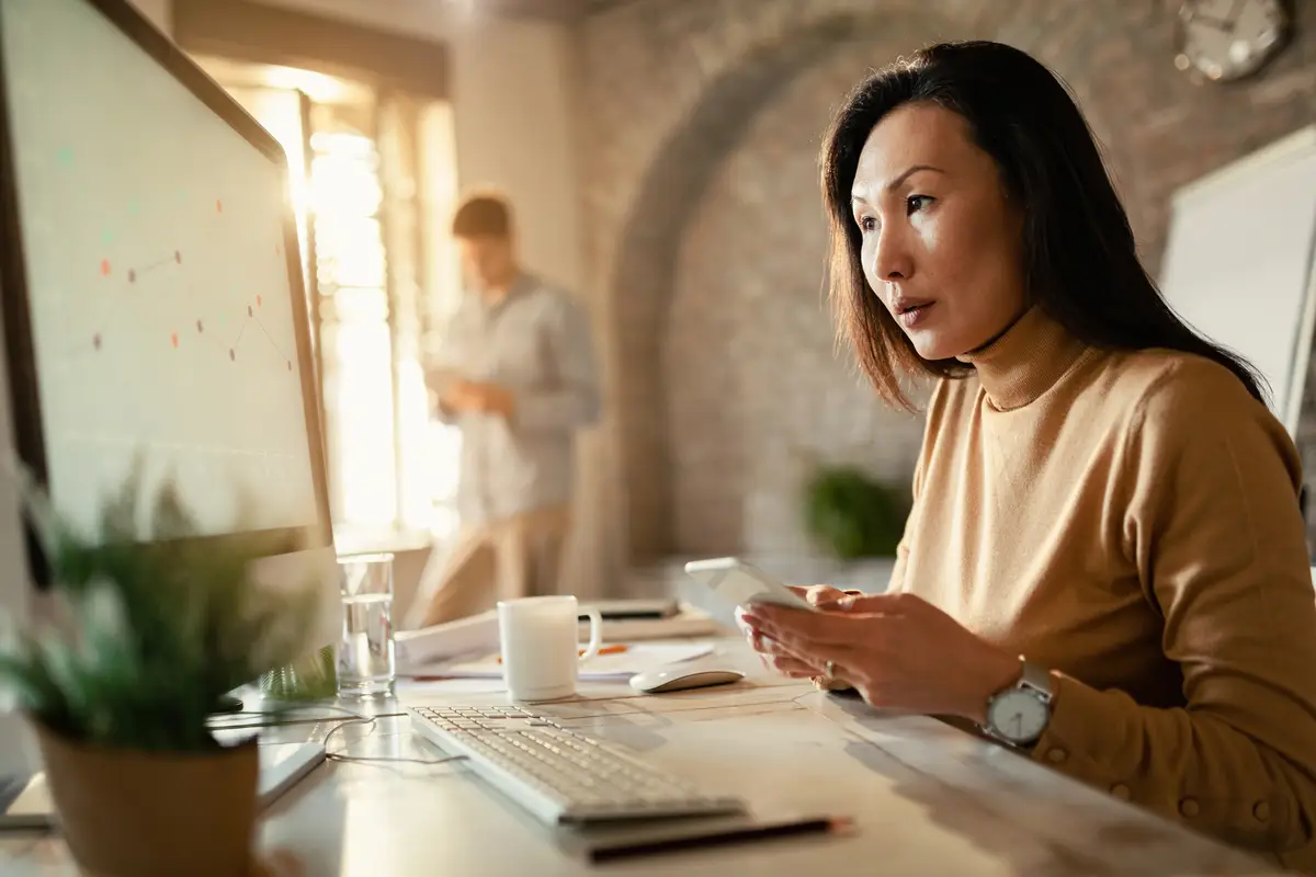 Female Asian entrepreneur reading charts on computer monitor and texting on smart phone in the office
