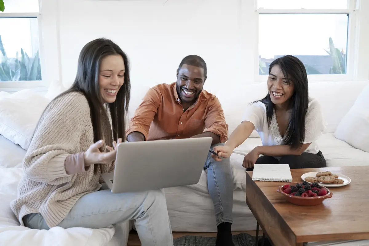 Group of young people using laptop together at home on sofa and having snacks