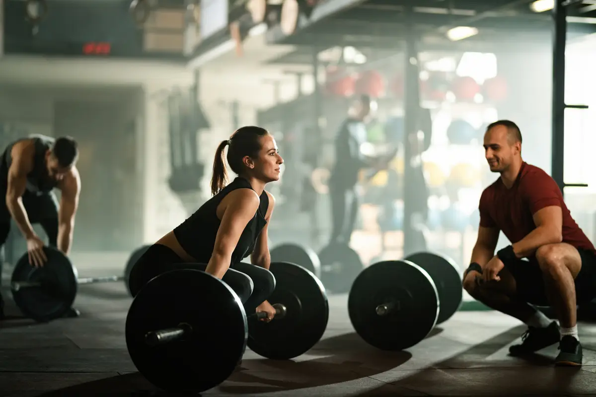 Young athletic woman lifting barbell while having strength training with her instructor in a gym