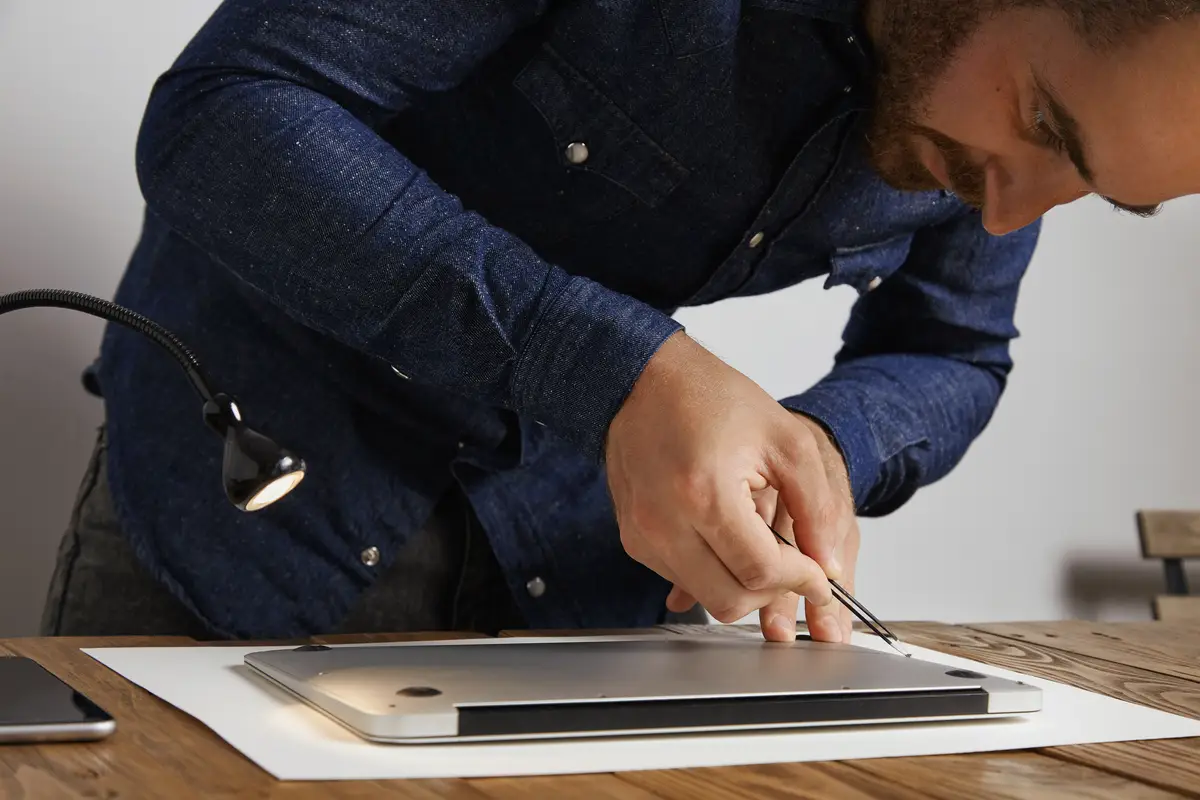 Serviceman puts small screws in hole with angled tweezers to close backside topcase of personal computer laptop after repairing and cleaning service in his lab