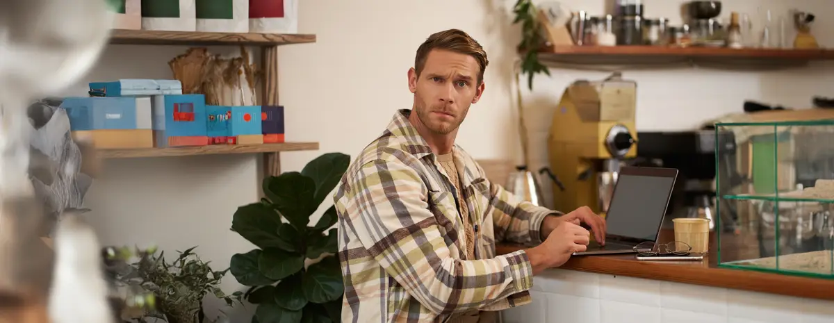 Portrait of man working in cafe looking confused at camera sitting with laptop and glass of coffee