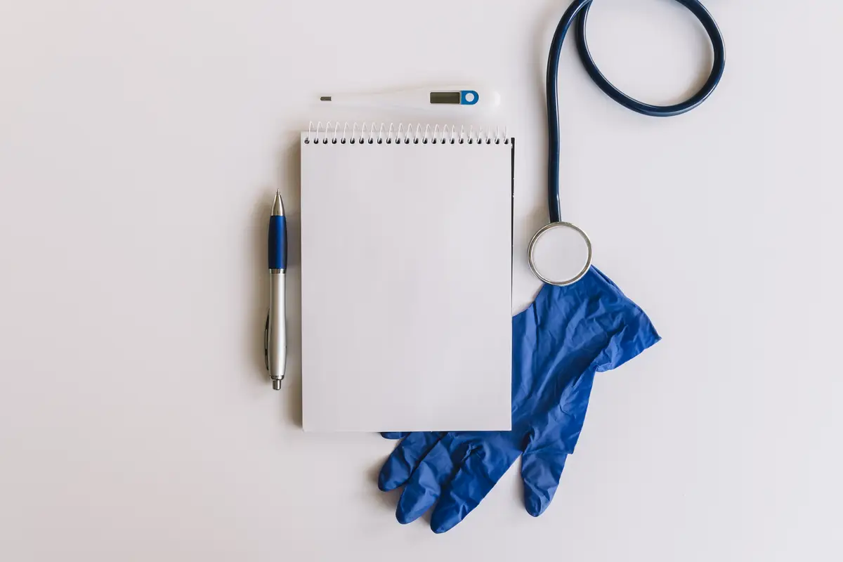 Spiral notepad; pen; stethoscope; thermometer and gloves on white surface