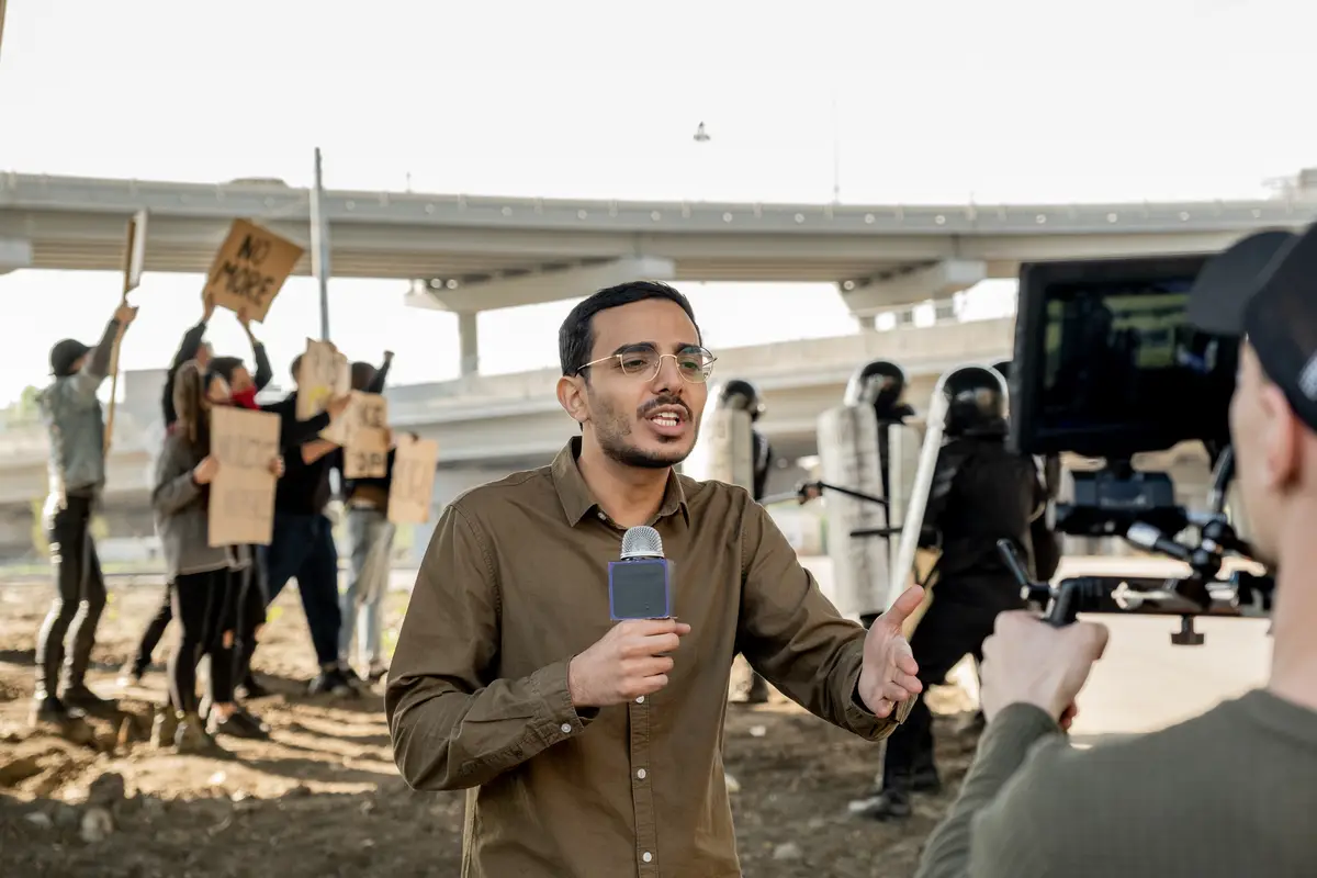 Young mixed race reporter with beard using microphone while covering riot in city: riot police keeping crowd of protestors