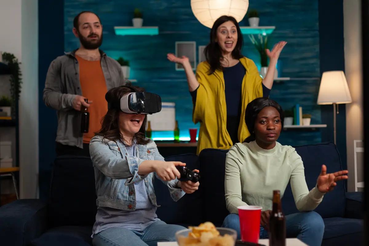 Happy gamer woman with virtual reality headset winning online competition playing videogames using gaming joystick enjoying victory with friends at home. Concept of multu-ethnic friends hanging out