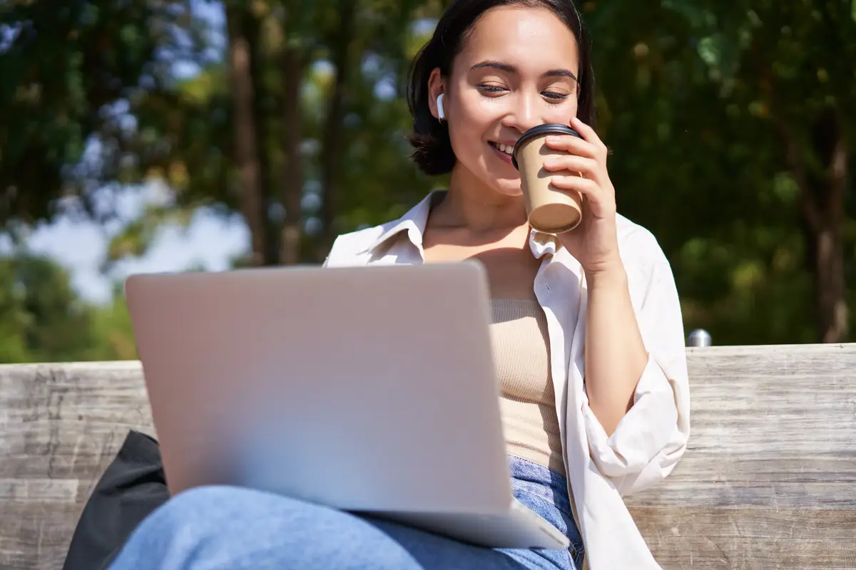 Asian girl sitting with laptop and wireless earphones drinking coffee looking at screen doing homewo