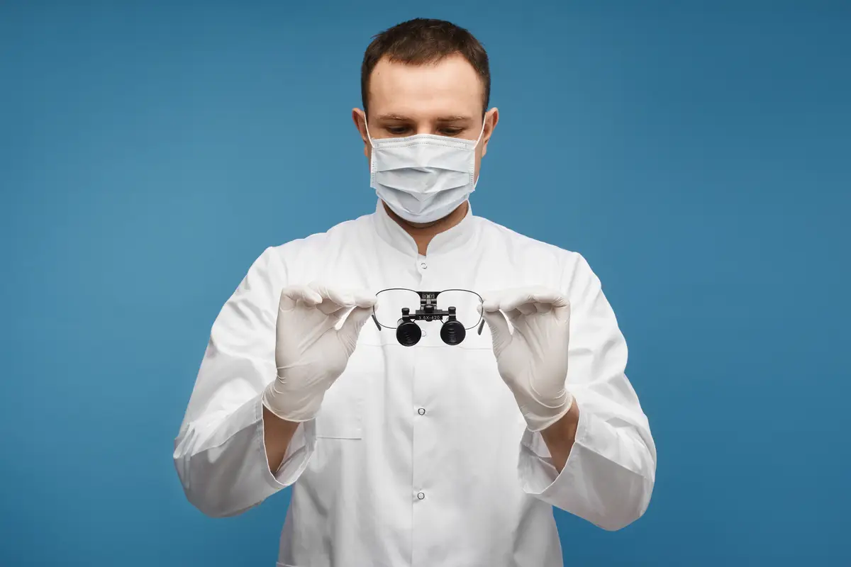 Male doctor in a surgical mask and protective gloves keeps binocular loupes in his hands, isolated at the blue background with copy space