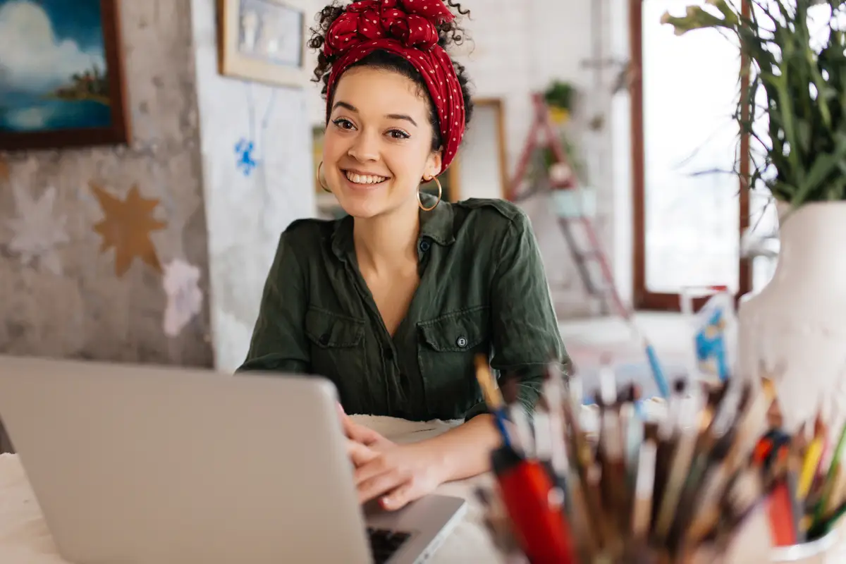 Young attractive woman with dark curly hair sitting at the table with laptop dreamily looking in camera while spending time in modern cozy workshop with big windows