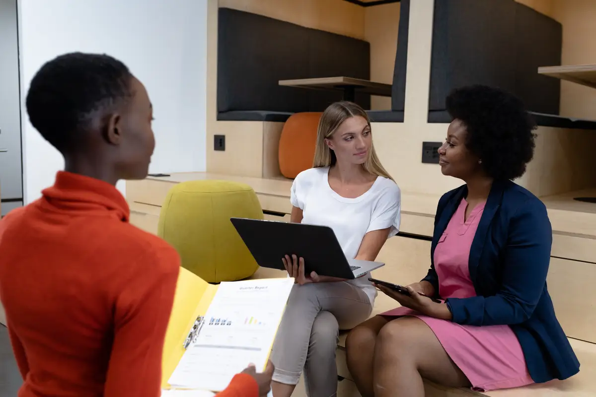 Diverse group of female business colleagues in discussion holding documents using tablet and laptop