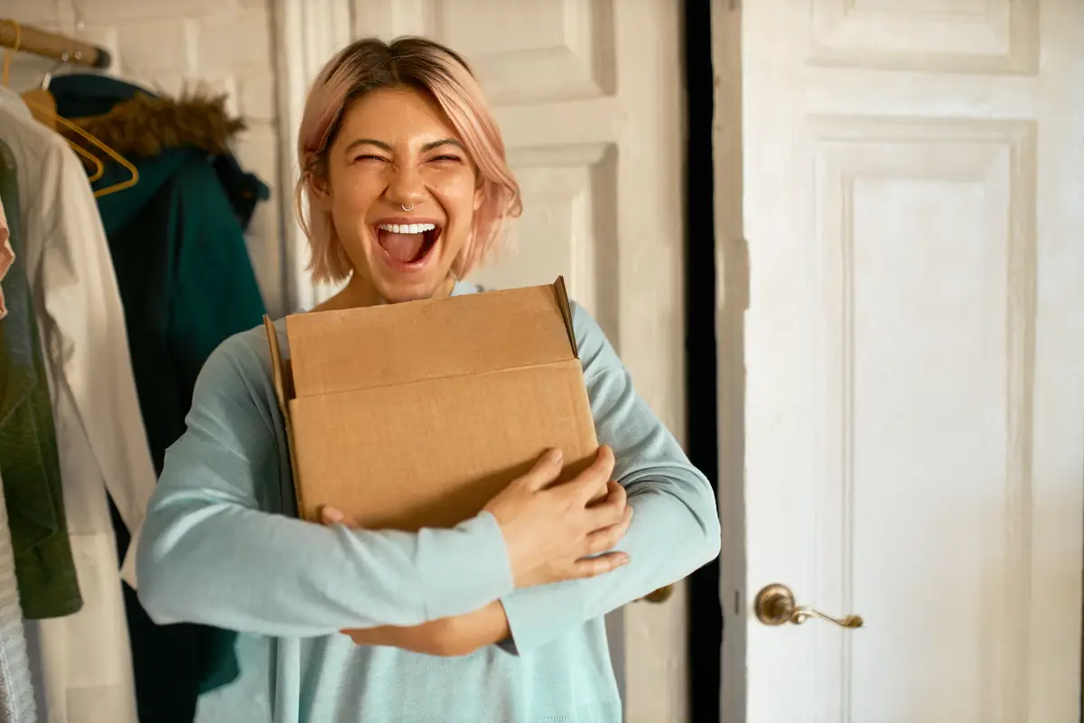 Indoor image of happy cheerful young woman holding cardboard box delivered to her apartment, expressing excitement, going to unpack parcel