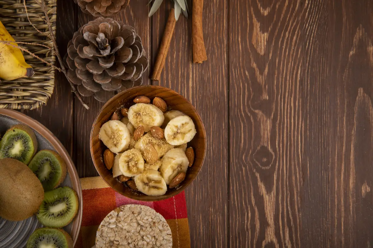 Top view of sliced bananas with almond in a wood bowl, slices of kiwi fruit on a plate and rice crackers on rustic with copy space