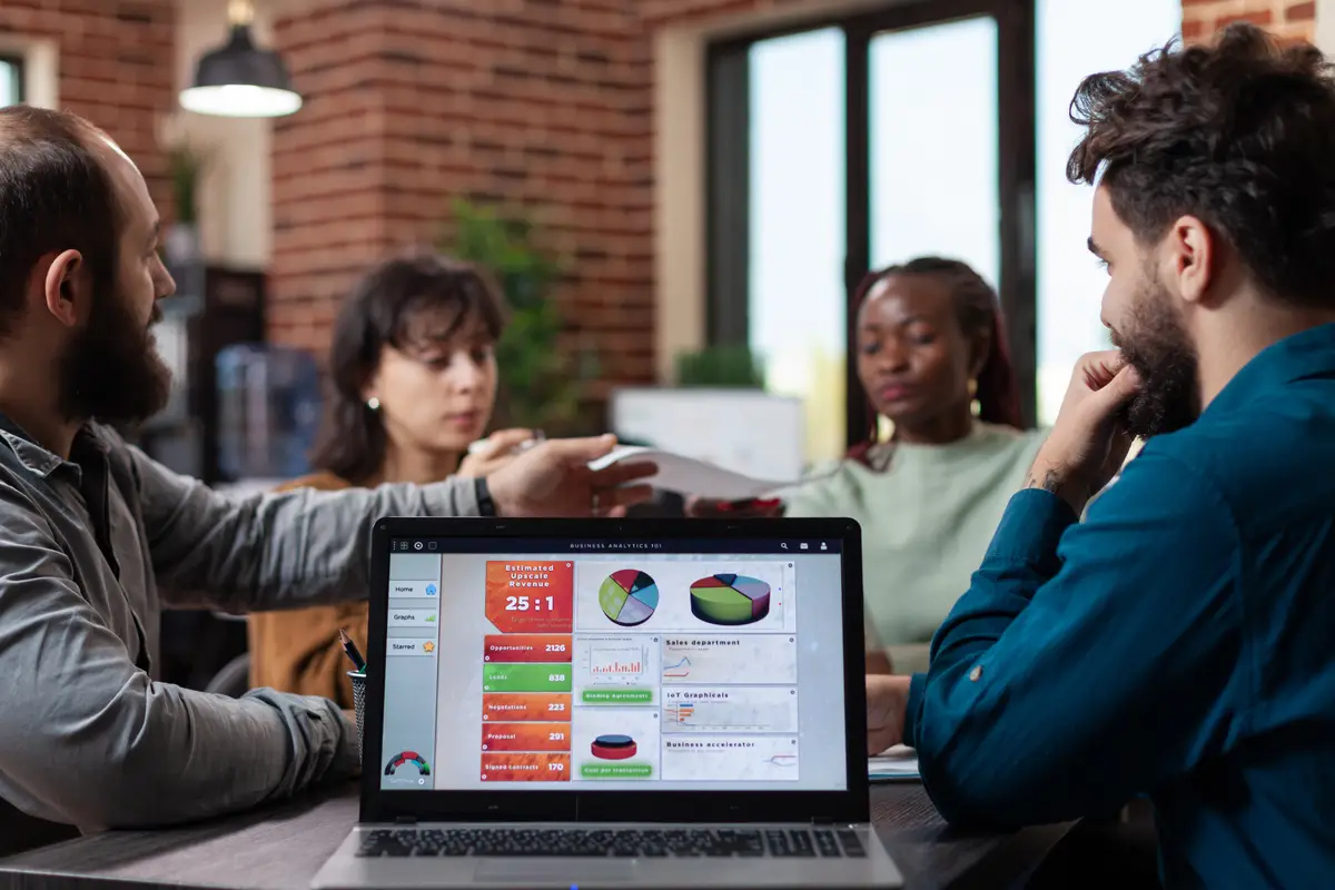 Laptop computer with marketing graphs on screen standing on table in startup office while diverse team sharing papers with company turnover on it. Multiethnic businesspeople analyzing strategy