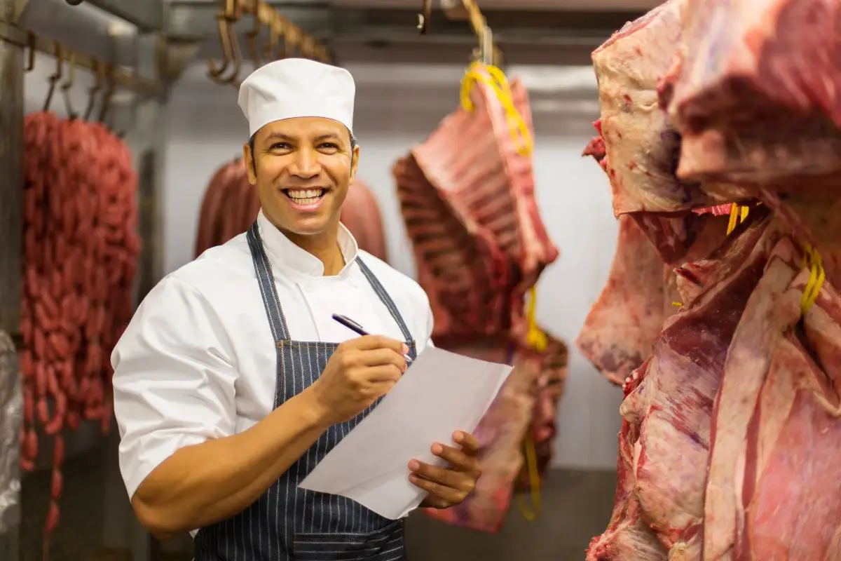 Açougueiro sorridente em câmara frigorífica segurando prancheta e caneta, com carne bovina pendurada ao redor
