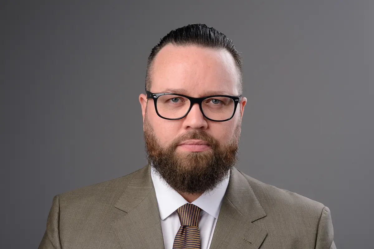 Homem sério de barba e óculos escuros vestindo terno marrom e gravata em fundo cinza