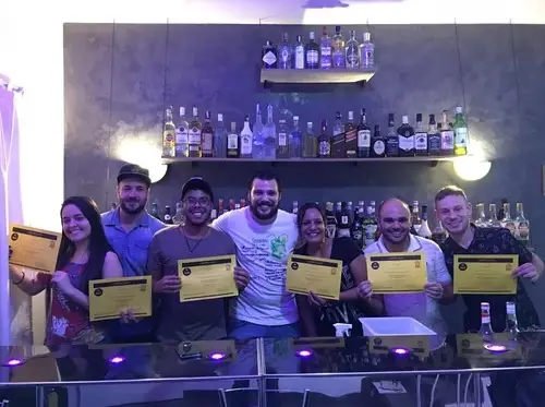 Seis pessoas segurando certificados em uma escola de bartenders com uma estante de bebidas ao fundo
