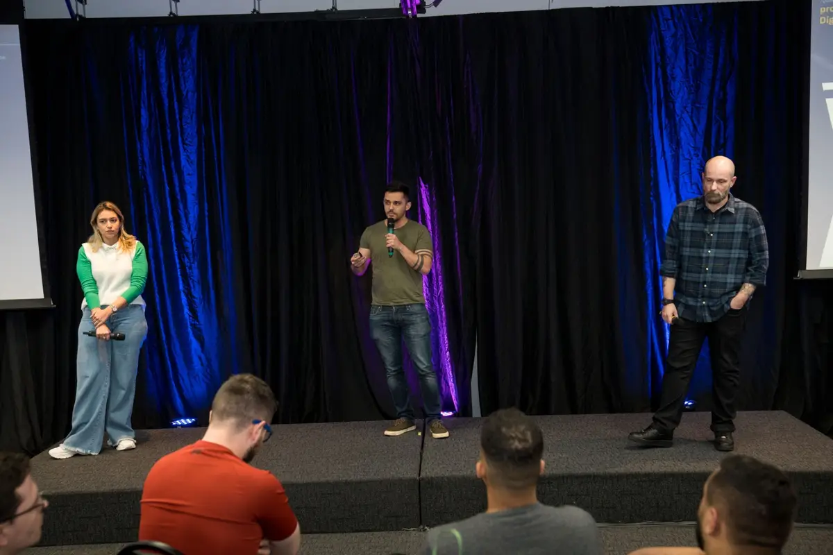 Três pessoas em um palco apresentando para uma audiência sentada, com cortinas pretas e iluminação azul e roxa ao fundo.