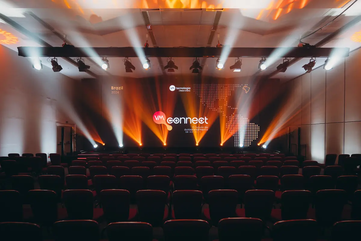Auditório com fileiras de cadeiras vazias voltadas para um palco iluminado em tons de laranja e branco com projeção de mapa e logo do evento VM Connect