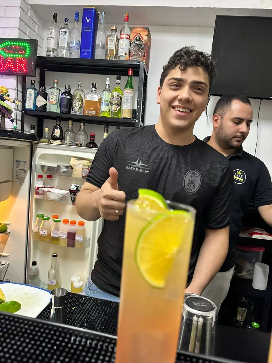 Bartender sorridente com camiseta preta fazendo gesto de positivo atrás do balcão de bar com coquetel decorado com fatias de limão em primeiro plano