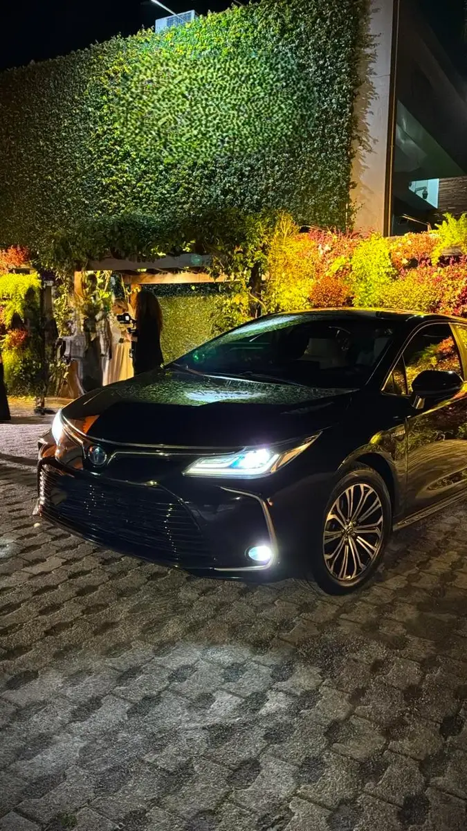 Sedã híbrido premium preto com faróis acesos estacionado em calçada de pedra à noite com parede verde iluminada ao fundo